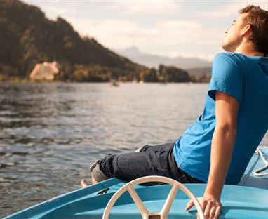 Ein junger Mann sitzt entspannt auf einem Boot und genießt die Aussicht auf das Wasser und die umliegenden Berge. Die Atmosphäre ist ruhig und friedlich.
