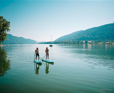 Zwei Personen stehen auf Stand-Up-Paddle-Boards auf einem ruhigen Gewässer. Im Hintergrund sind Hügel und ein klarer, blauer Himmel zu sehen.