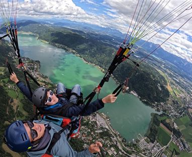 Zwei Paraglider schwebend über einer grünen Landschaft mit Seen und Bergen. Der Himmel ist teilweise bewölkt und bietet einen spektakulären Ausblick.