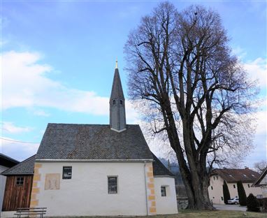 Eine kleine Kirche mit einem spitzen Turm steht neben einem großen Baum. Der Himmel ist blau mit einigen Wolken.