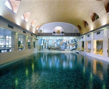 Ein stilvolles Hallenbad mit klarem Wasser und einem hohen, architektonisch ansprechenden Deckenbereich. Die Wände sind mit künstlerischen Gemälden dekoriert.