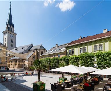 Ein schöner Platz mit Cafés und Sonnenschirmen. Im Hintergrund ist ein Kirchturm und eine grüne Fassadenlandschaft zu sehen.