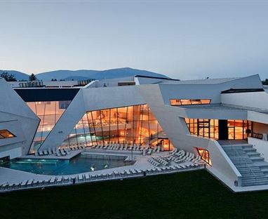 Ein modernes, architektonisch interessantes Haus mit großen Fenstern und einem Swimmingpool. Die Umgebung ist grün und bietet eine malerische Aussicht auf die Berge.