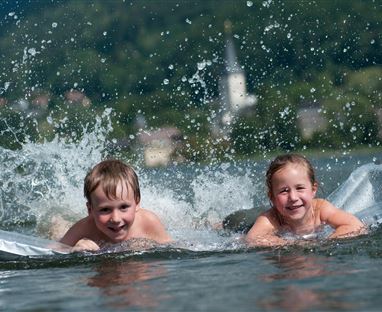Zwei Kinder schwimmen fröhlich im Wasser und spritzen dabei. Im Hintergrund ist eine malerische Landschaft mit einem Kirchturm zu sehen.