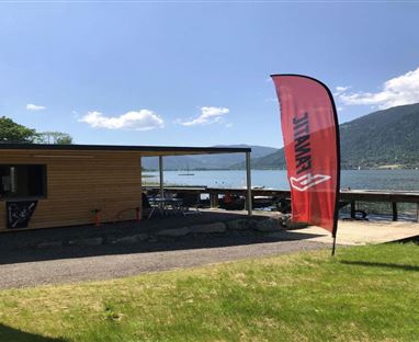 Ein gemütliches Holzhaus am Ufer eines Sees, umgeben von grüner Wiese. Ein rotes Werbebanner weht im Wind, während die Berge im Hintergrund zu sehen sind.