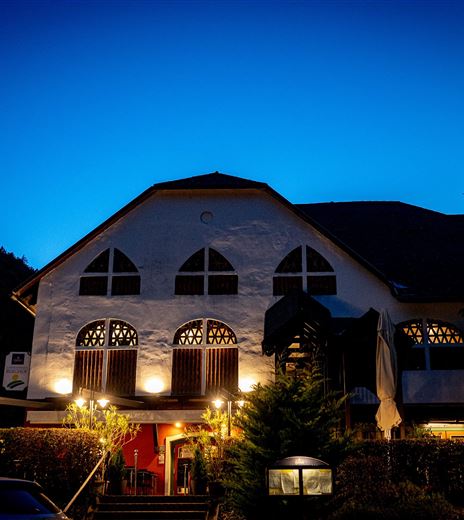 Ein charmantes Gebäude bei Nacht, beleuchtet von warmem Licht. Der Himmel ist klar und dunkelblau, umgeben von Bäumen.