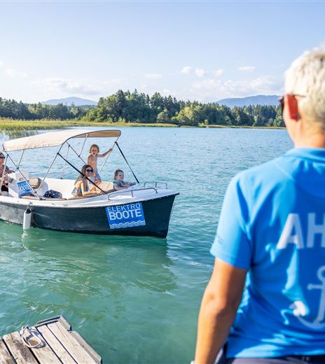 Ein Boot mit mehreren Personen fährt auf einem blauen See. Im Vordergrund steht eine Person mit einem blauen T-Shirt, das "AHOI!" zeigt.