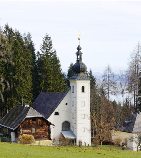 Eine malerische Kirche mit einem hohen Turm, umgeben von grünen Wäldern. Im Hintergrund sind sanfte Hügel und Bäume sichtbar.
