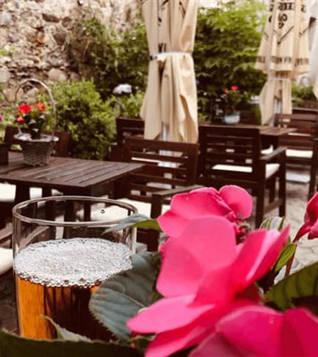 Ein gemütlicher Innenhof mit Holztischen und Stühlen. Im Vordergrund steht ein Glas Bier neben pinken Blumen.