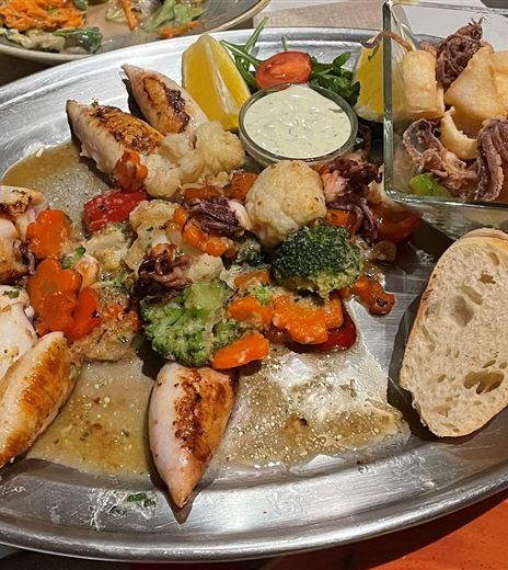 Un grande piatto di portata con frutti di mare alla griglia, verdure e pane. Sono incluse due ciotole con salse e ulteriori frutti di mare.