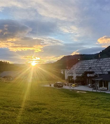 Eine malerische Landschaft mit einem Sonnenuntergang hinter einem Holzhaus. Im Vordergrund erstreckt sich eine grüne Wiese unter einem bewölkten Himmel.