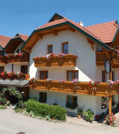 Ein gemütliches Hotel im traditionellen Stil mit Holzverzierungen und Balkonen voller Blumen. Die Sonne scheint auf die farbenfrohe Fassade und die umliegende grüne Landschaft.
