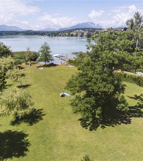 Eine weitläufige Grünfläche mit mehreren Bäumen und Blick auf einen ruhigen See. Im Hintergrund sind Berge und einige Häuser sichtbar.