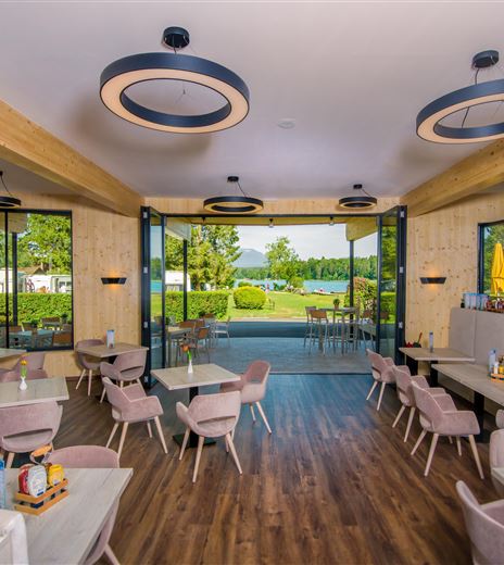 Ein modernes Restaurant mit gemütlichen Tischen und Stühlen. Große Fenster bieten einen Blick auf die grüne Landschaft draußen.
