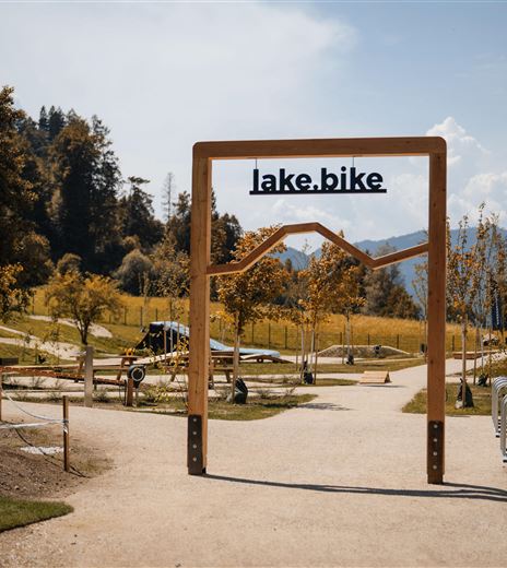 An access sign for lake.bike in a scenic environment. The path leads through a park with trees and recreational opportunities.