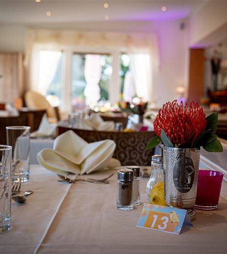 Una tavola elegantemente apparecchiata in un ristorante con fiori e elementi decorativi. Sullo sfondo si vede una cameriera che prepara il servizio.