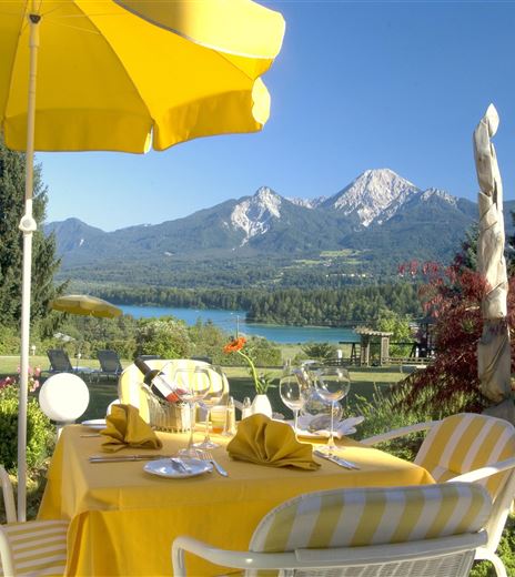 Ein Tisch mit gelben Servietten und einem Sonnenschirm steht in einem Garten mit Blick auf die Berge und einen See. Die Umgebung ist malerisch und einladend.