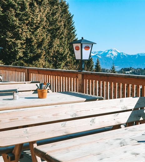 Un'accogliente area esterna con tavoli e panche di legno. Sullo sfondo si ergono maestose montagne sotto un chiaro cielo blu.
