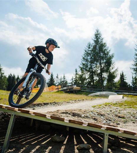 Ein Junge fährt mit seinem Mountainbike über eine Rampe. Im Hintergrund sieht man Bäume und einen klaren Himmel.