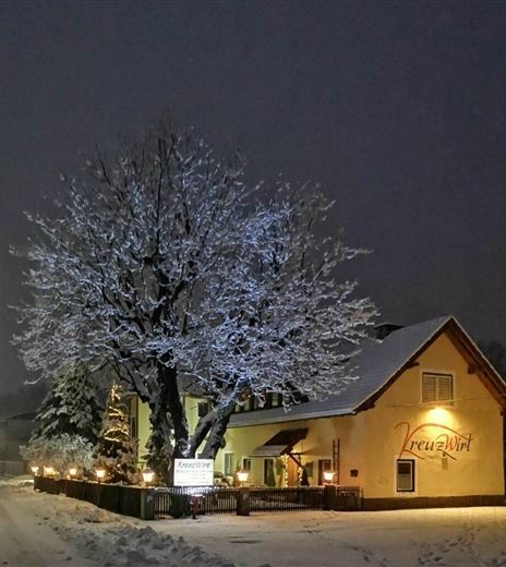 Ein gemütliches Gasthaus mit einem hell erleuchteten Schild bei Nacht. Der schneebedeckte Baum und die Umgebung tragen zur winterlichen Atmosphäre bei.