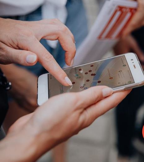 Eine Hand hält ein Smartphone, auf dem eine Kartenansicht angezeigt wird. Im Hintergrund sind Personen und eine Informationsbroschüre zu sehen.