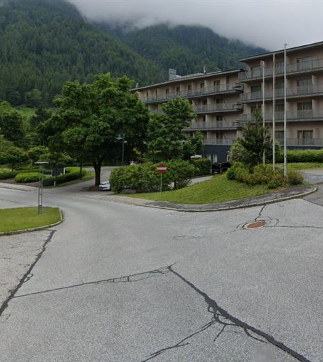 Ein moderner Gebäudekomplex umgeben von Bäumen und Bergen. Im Vordergrund ist eine Straße mit parkenden Autos zu sehen.