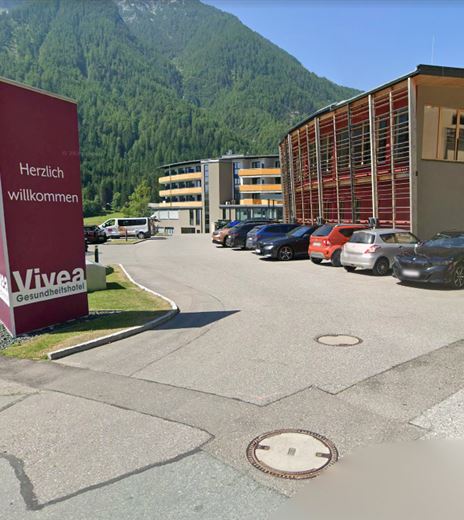 Ein Eingang zu einem Gesundheitszentrum mit einem großen Willkommensschild. Im Hintergrund sind mehrere Autos und eine bergige Landschaft zu sehen.