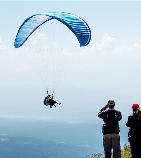 Ein Paraglider schwebt über einer schönen Berglandschaft. Zwei Zuschauer beobachten das Geschehen gespannt.