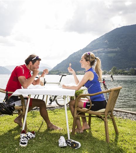Ein Paar sitzt am Tisch am Ufer eines Sees. Sie genießen eine entspannte Mahlzeit mit Blick auf die Landschaft und die Berge.