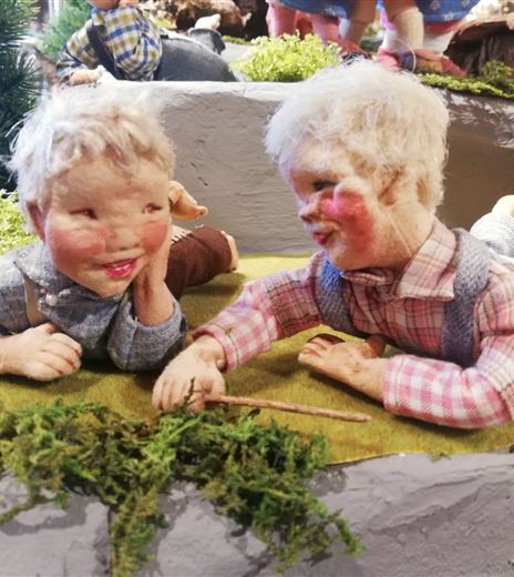 Zwei Keramikkinder spielen fröhlich auf einer Wiese. Sie liegen nebeneinander und scheinen einander etwas zu erzählen.
