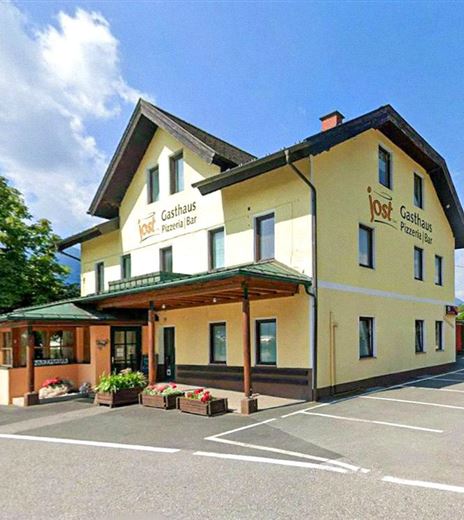 An inn with a light yellow facade and a green roof. In front of the building, there is a parking lot and some plants.