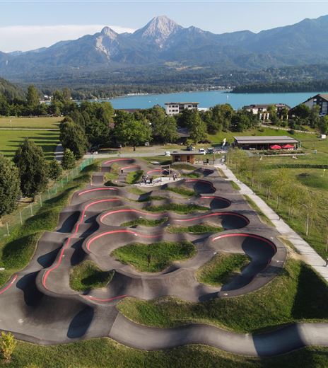 Ein moderner Pumptrack in einer grünen Landschaft mit Bergen im Hintergrund. Der See glitzert in der Sonne, während Menschen aktiv auf dem Track sind.