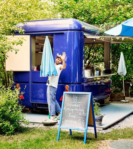 Un accogliente food truck nella natura, circondato da alberi. Un uomo amichevole saluta davanti al truck, mentre viene posizionato un ombrello.
