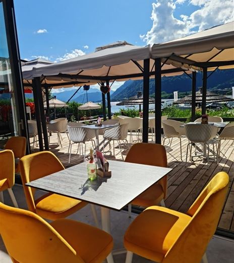 Ein modernes Café mit orangefarbenen Stühlen und einem Tisch im Vordergrund. Im Hintergrund sind Sonnenschirme und eine schöne Landschaft mit Bergen zu sehen.