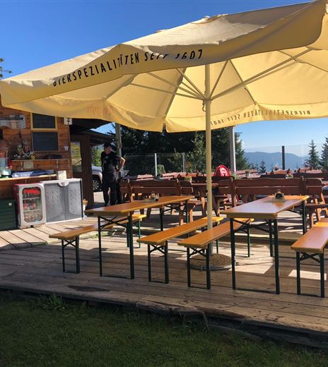 Eine rustikale Terrasse mit Holzbänken und Tischen unter einem großen Sonnenschirm. Im Hintergrund steht eine kleine Hütte in einer grünen Umgebung.