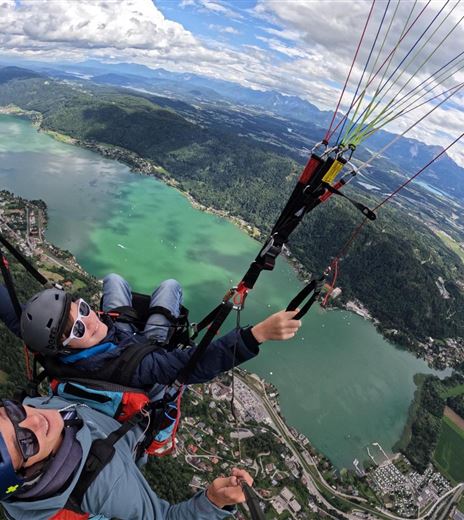 Two paragliders hovering over a green landscape with lakes and mountains. The sky is partially cloudy and offers a spectacular view.