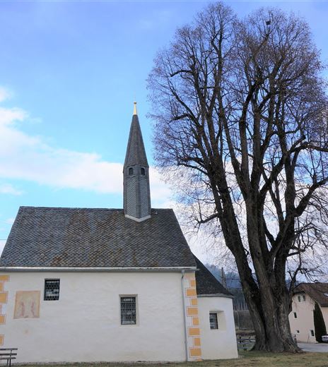 Eine kleine Kirche mit einem spitzen Turm steht neben einem großen Baum. Der Himmel ist blau mit einigen Wolken.