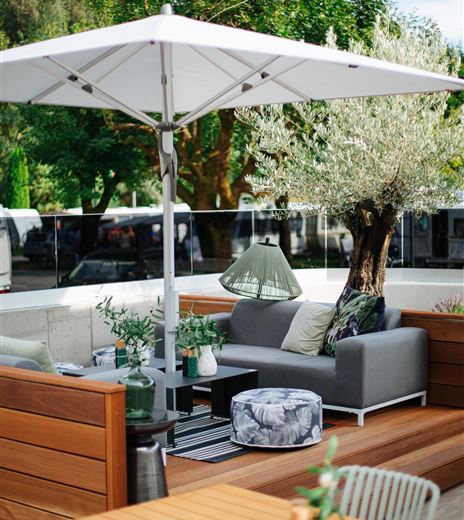 Un'accogliente area esterna con un divano grigio, un grande ombrello da sole e piante ben curate. L'ambiente è arredato in modo moderno e invita a rilassarsi.