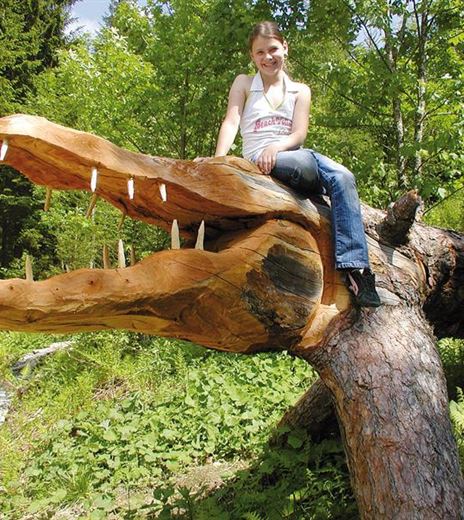 Una ragazza è seduta sulla testa di una grande figura in legno di coccodrillo, finemente intagliata. Sullo sfondo si vede un paesaggio verdeggiante e boscoso.