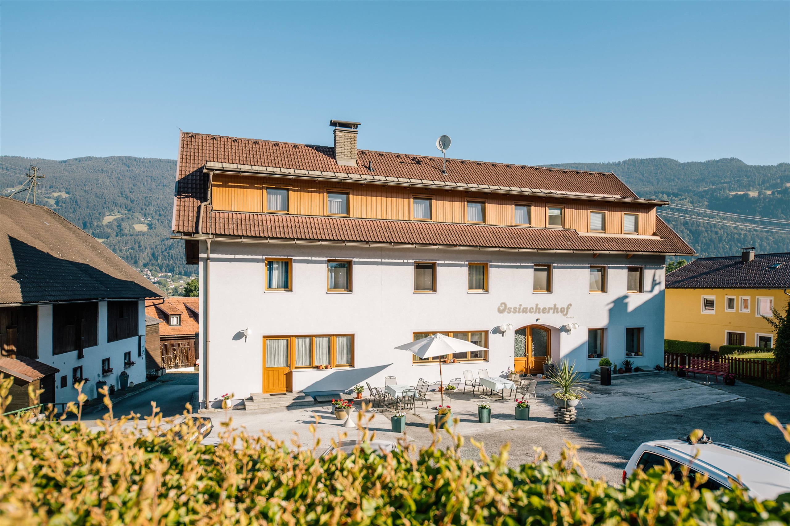 Ein gemütliches Gasthaus in einer malerischen Berglandschaft. Die Fassade ist weiß mit Holzverkleidungen und es gibt eine überdachte Terrasse.