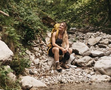 A woman is sitting on stones next to a clear stream in a forest. She is wearing a backpack and appears relaxed in nature.