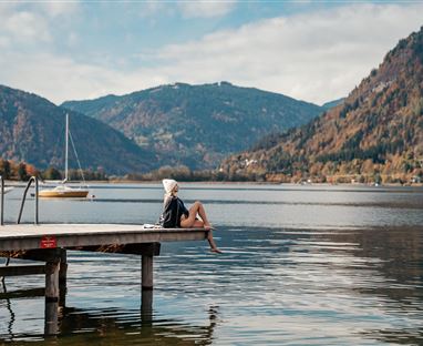 Una persona è seduta sulla riva di un lago tranquillo e guarda le montagne circostanti. Il paesaggio è idilliaco con dolci colline e barche sullo sfondo.