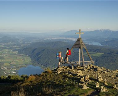 Zwei Wanderer stehen an einer Aussichtsplattform mit einem Holzkreuz auf einem Berggipfel. Im Hintergrund sind weite Täler und Berge mit klarem Himmel zu sehen.