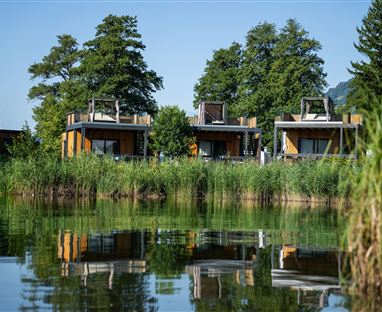 Modern vacation homes on the shore of a clear lake. The buildings are surrounded by lush greenery and reflect in the water.