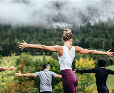 Eine Gruppe von Menschen macht Yoga in der Natur. Im Hintergrund sind Berge und grüne Wälder zu sehen.
