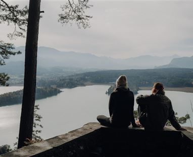 Due persone sono sedute su un promontorio di pietra e guardano un lago tranquillo. Sullo sfondo si estendono dolci colline e boschi.