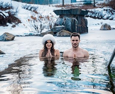 A couple stands in an icy water basin in a snowy environment. The woman is wearing a white fur coat and both are showing a meditation gesture.