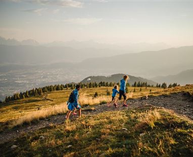 Eine Gruppe Kinder wandert auf einem Bergpfad. Im Hintergrund ist eine atemberaubende Landschaft mit schneebedeckten Bergen und einem klaren Himmel zu sehen.