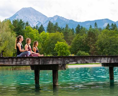 Three women are meditating on a wooden walkway over clear water. In the background, mountains and green trees can be seen.
