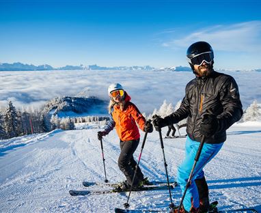 Zwei Skifahrer stehen auf einer schneebedeckten Piste mit herrlichem Ausblick auf Wolken und Berge. Der Himmel ist klar und blau, ideal für einen Tag im Schnee.